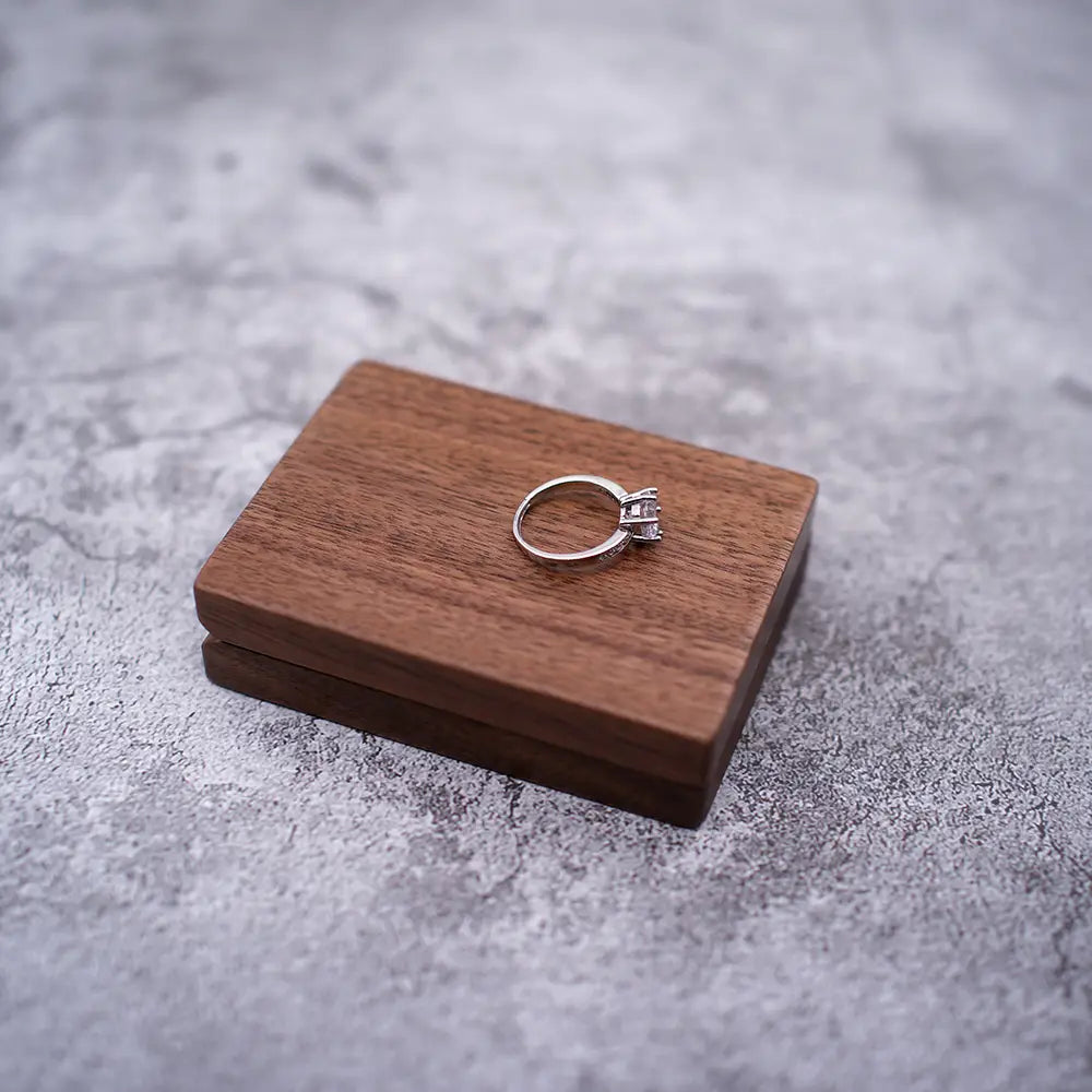 Ein silberfarbener Verlobungsring mit einem zentralen Diamanten und kleineren Akzentsteinen auf einem eleganten, minimalistischen Band, das auf einer personalisierten Schmuckring-Schachtel aus Walnussholz zur Hochzeit ruht.
