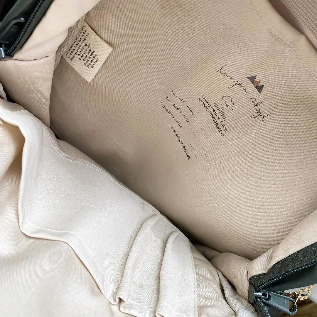 Beige Innenfutter-Auskleidung eines puffer jacket von Konges sløjd für Kinder mit besticktem Markenlogo und Pflegehinweisen in einem gesteppt bestickten, wasserfesten Kinder-Schulrucksack für den Schulgebrauch