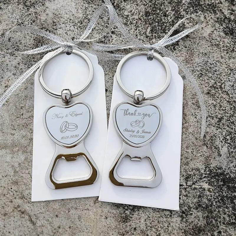 Porte-clés décapsuleurs en métal argenté en forme de cœur avec gravure des détails du mariage et anneaux fixés à des étiquettes blanches avec un ruban argenté, pour des faveurs de mariage personnalisées en décapsuleur en forme de cœur.