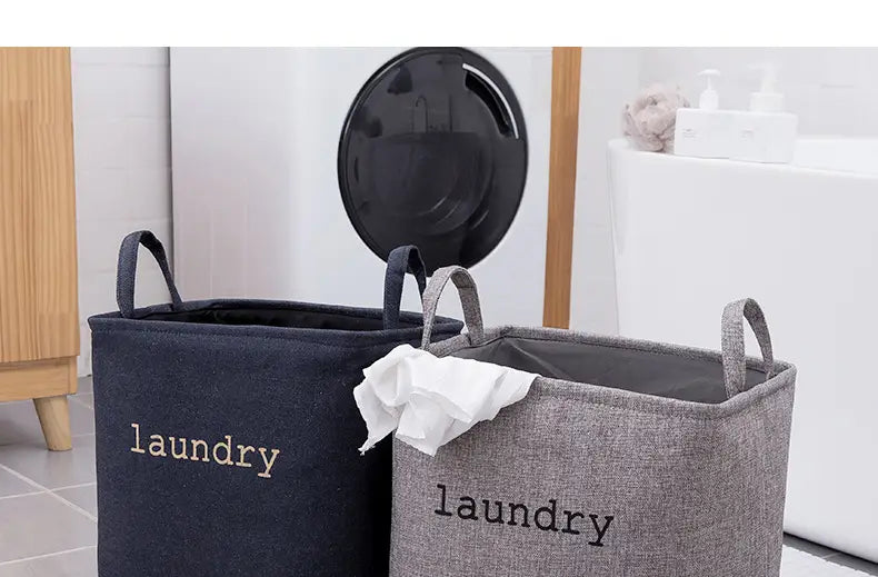 two foldable laundry baskets made of fabric in dark gray and light gray with black handles and laundry embroidered on them for use as storage baskets.
