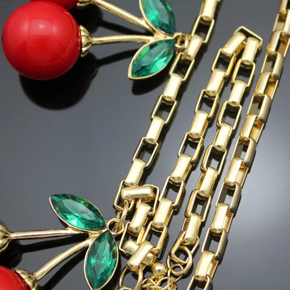 Un collier à chaîne à tonalité dorée, orné de charmes de cerises rouges et émeraude éclatants et de feuilles en pierre serties facettées, le tout dans un collier statement de cerises rouges avec collier ras du cou en cristal.