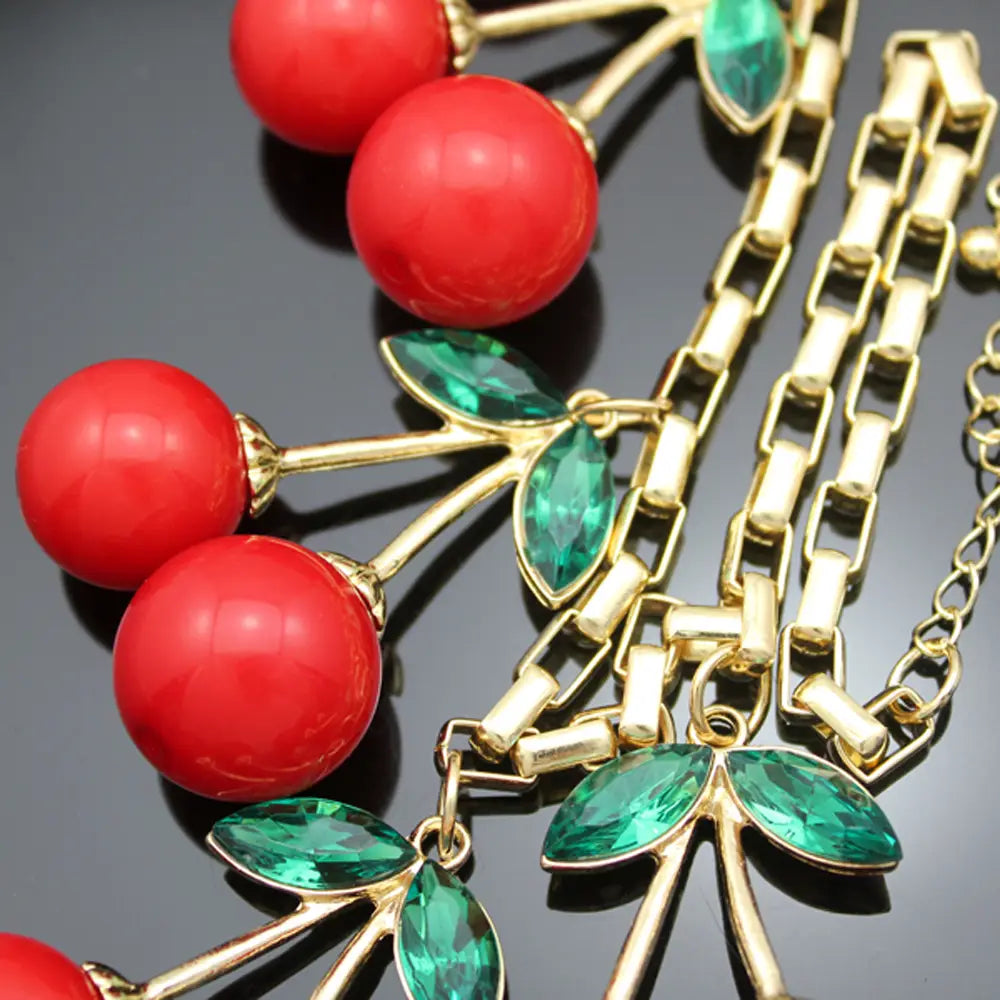 Un collier statement de cerises rouges à tonalité dorée, mettant en valeur des charmes sphériques rouges éclatants et des accents en cristal de feuille verte facettée.