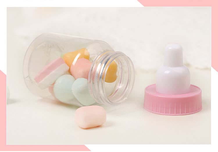 ピンクのねじ込み式キャップと白い乳首を備えた透明なプラスチック製ベビーシャワー引き菓子用ミニミルクボトル。横向きに置かれており、パステルカラーの入浴用ボムを入れた開いた瓶のそばにあります。