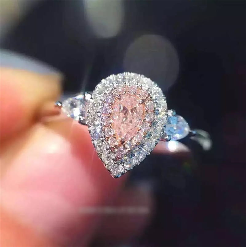 Bague de fiançailles délicate en zircon goutte de larme rose à monture argentée, présentant un diamant rose central en forme de poire entouré d’un halo de diamants transparents, rehaussé de petites pierres taillées rondes de chaque côté.