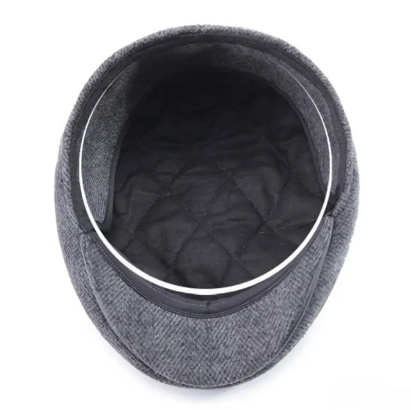 Gorra plana tipo herringbone para hombre confeccionada en lana gris, con interior acolchado negro y ribete plateado, con cubreorejas para uso en invierno.