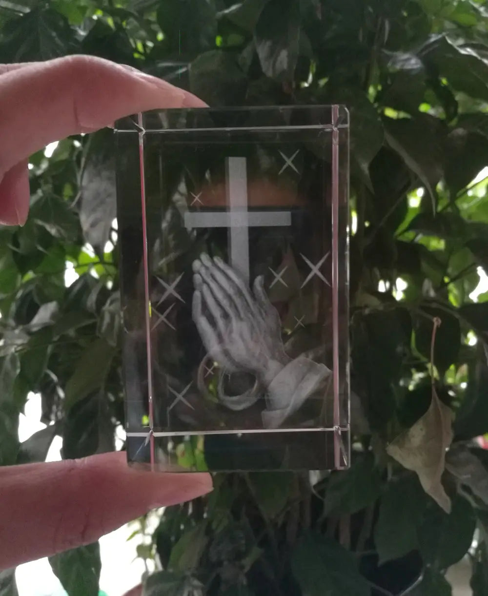 Cube en cristal transparent 3D en forme de croix avec mains priantes, avec une croix blanche gravée et un motif de mains priantes.