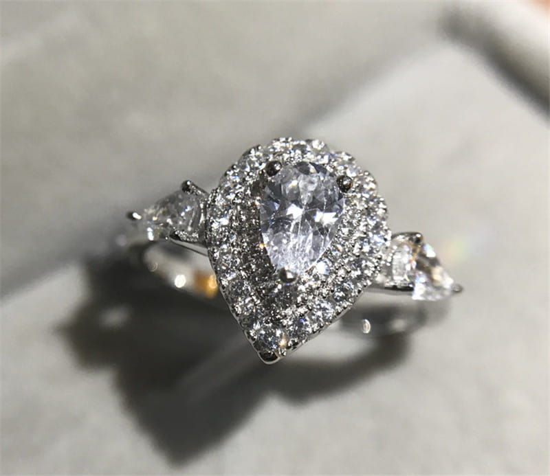 Bague de fiançailles en zircon goutte de larme rose en forme de poire étincelante, avec un halo de diamants plus petits et des pierres d’accent serties dans une bande métallique élégante à tons argentés.