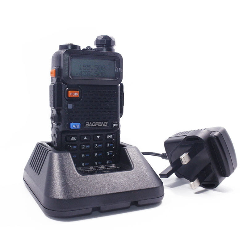 Black baofeng uv-5r handheld two-way radio with digital display and keypad resting on its charging base next to a black uk-style power adapter for baofeng uv-5r battery charger.