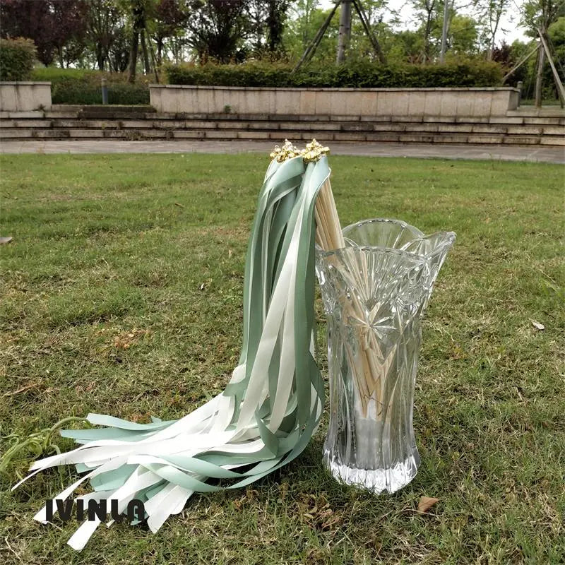 un bouquet délicat de baguettes ruban de mariage vert clair et blanc, avec des accents floraux dorés, disposé avec élégance à côté d’un vase en cristal transparent.