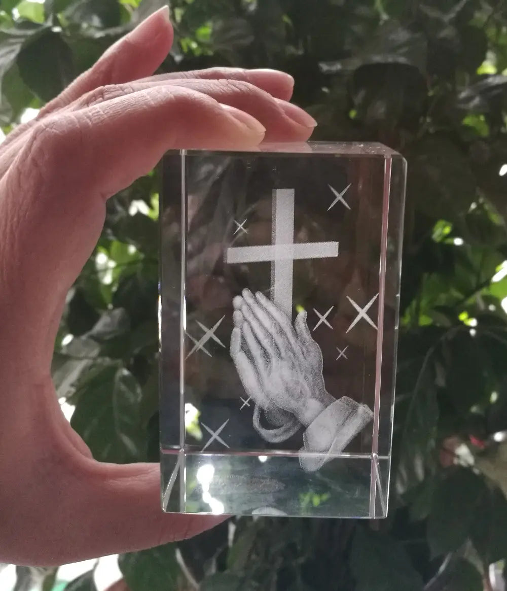 Bloc de cristal transparent rectangulaire présentant un motif finement gravé de mains priantes et une croix entourée de motifs semblables à des étoiles, dans une statue de croix en cristal 3D avec mains priantes.