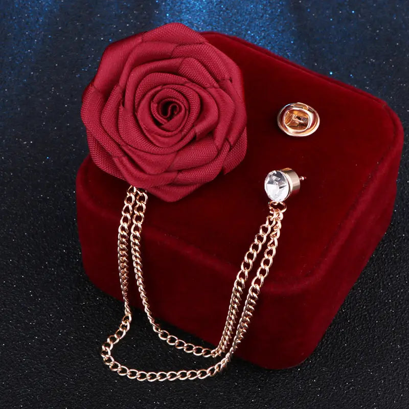 A deep red fabric rose brooch with a gold-toned tassel chain and crystal accent designed as a men suit pin.