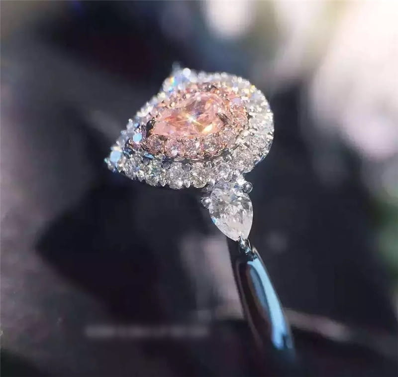 Bague de fiançailles délicate en zircon goutte de larme rose, présentant une pierre rose centrale en forme de poire entourée d’un halo de diamants transparents, avec un diamant pendentif taillé en goutte.