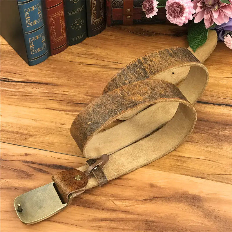A vintage-style brown leather men jeans belt with a brass owl belt buckle and visible wear resting on a wooden surface.