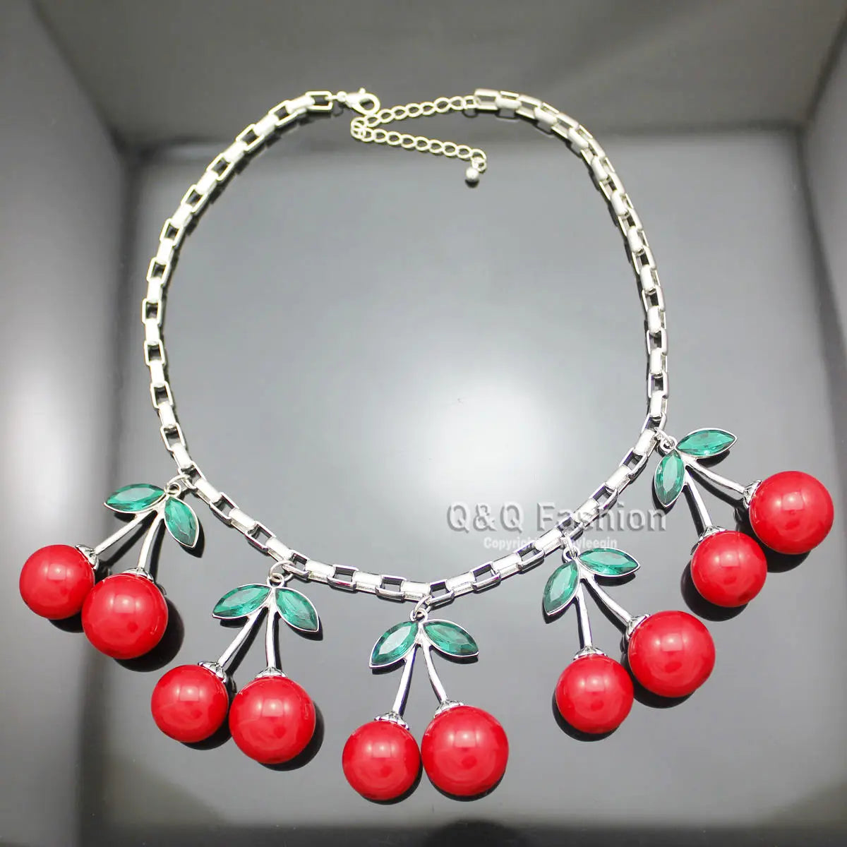 Un collier à chaîne argentée, composé de pendants cerises rouges éclatants, rehaussés d’accents de feuilles vertes, conçu comme un collier ras du cou en cristal statement de cerises rouges.
