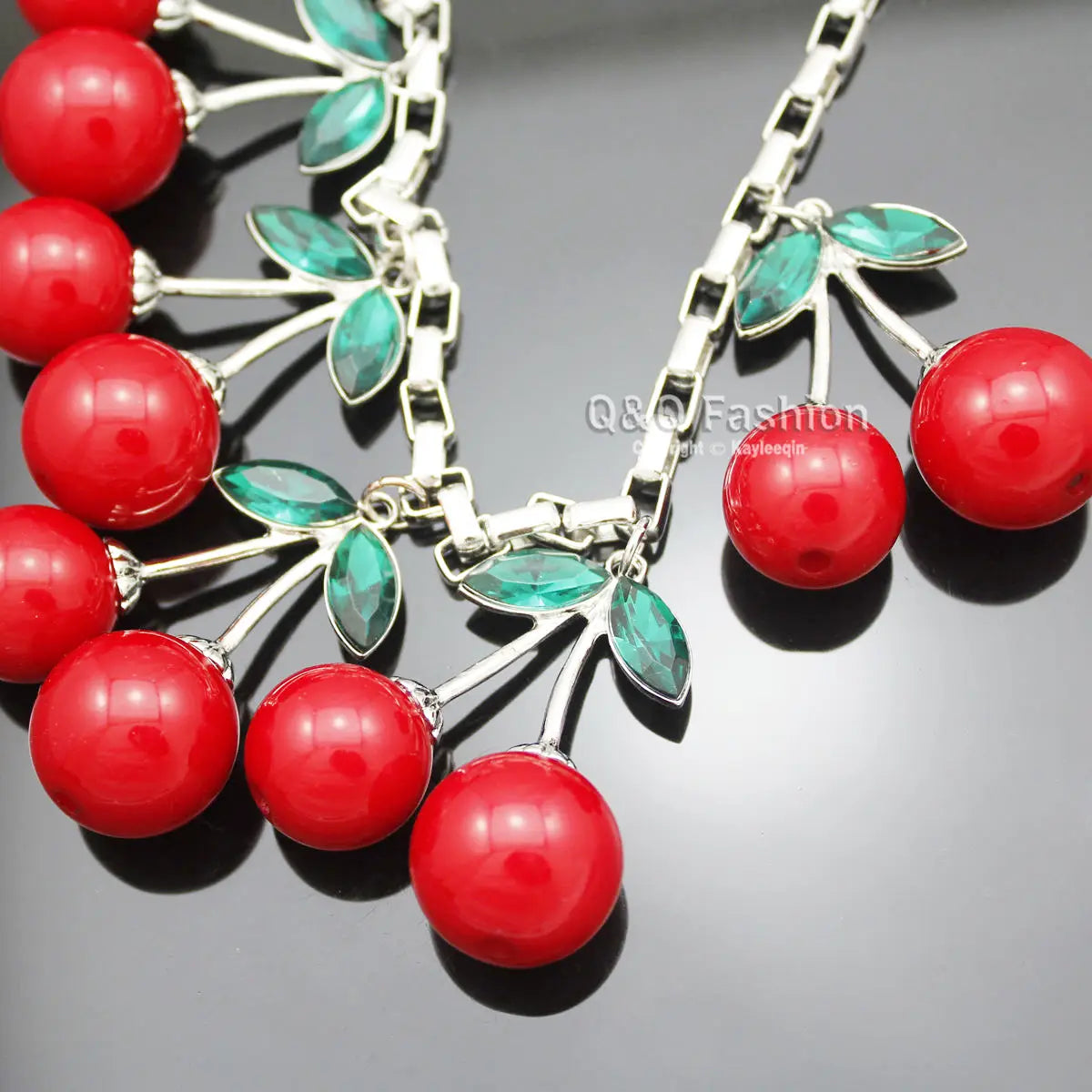 Un collier statement de cerises rouges éclatant, doté de cerises rouges en résine brillante et de feuilles vertes facettées sur une chaîne de collier ras du cou en cristal à tonalité argentée.
