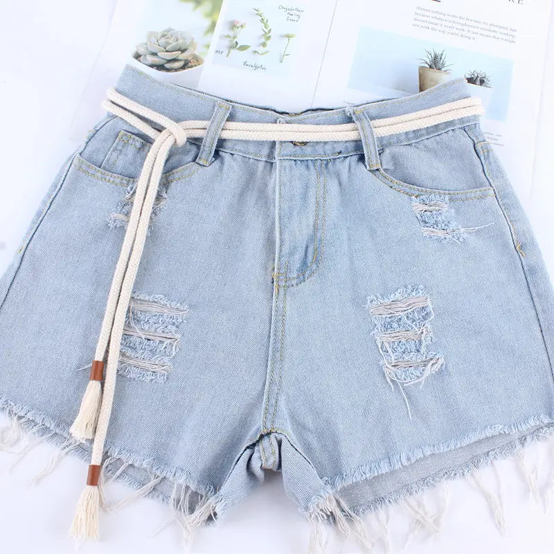 light-wash denim shorts with distressed rips and frayed hems accented by a cream-colored braided rope belt with tassels tied at the waist