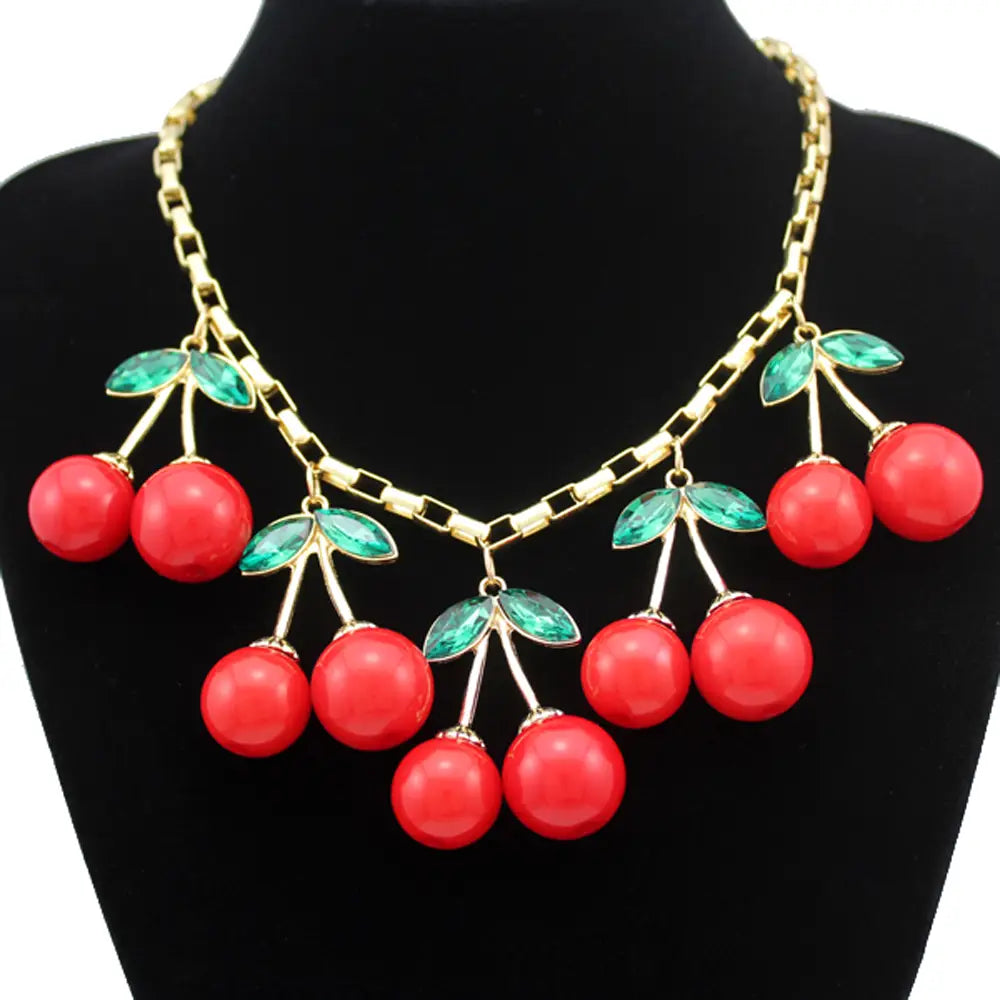 Un collier statement cerises rouges éclatant, avec un design de collier ras du cou en cristal, des chaînes à tonalité dorée et des touches de feuilles vertes.