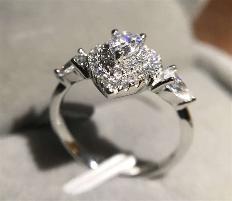 Bague de fiançailles en zircon goutte de larme rose étincelante à monture argentée, présentant un diamant central rond entouré d’un halo de diamants plus petits et de deux pierres d’accent triangulaires de part et d’autre.
