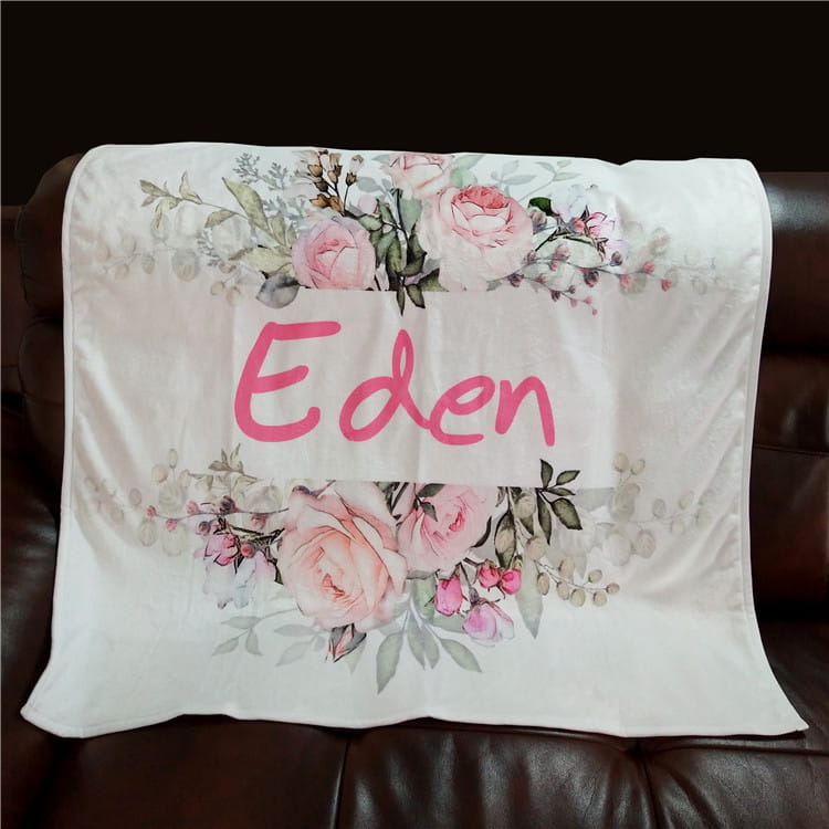 Eine weiße, personalisierte Baby-Wickeldecke aus Fleece mit einer zarten floralen Musterung aus pinken Rosen und grünen Blättern, mit dem Namen Eden in kräftiger pinkfarbener Schrift.