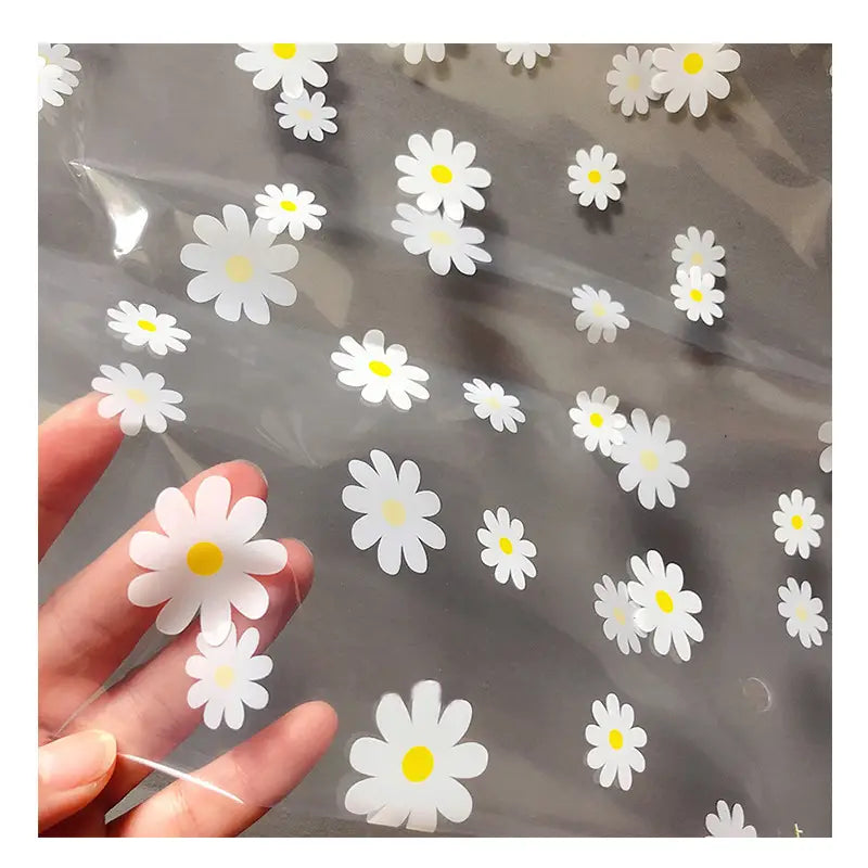 Gänseblümchenförmige Aufkleber mit gelben Zentren und einer glänzenden Oberfläche auf einer blumigen, durchsichtigen Reisekosmetiktasche.