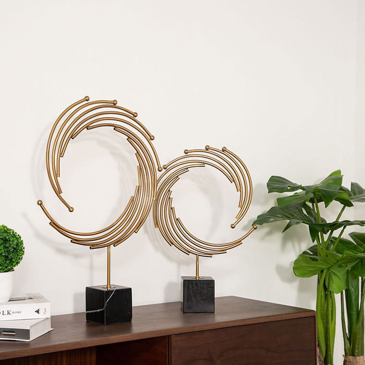Two gold-toned metal abstract sculptures with concentric circular designs on black marble bases for office desk decor.