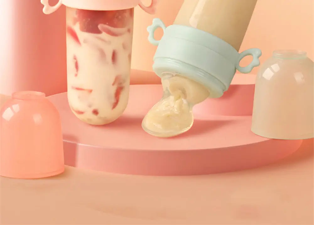 Pastellblaue und rosafarbene Silikon-Babysqueeze-Feedingflasche und Löffelspender mit einem klaren zylindrischen Körper, gefüllt mit Joghurt- und Erdbeerstücken.