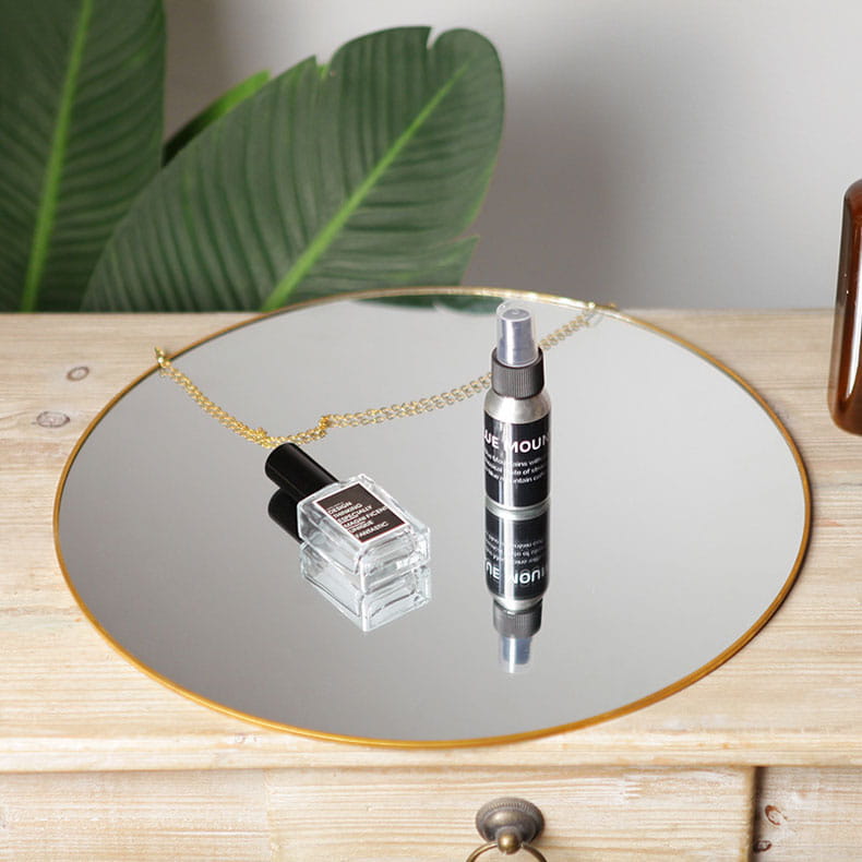 silver spray bottle with black label and transparent nozzle resting on a round gold hexagonal wall hanging mirror with gold frame alongside a clear glass perfume bottle