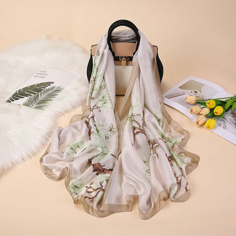 Un foulard en soie couleur crème délicat, avec un imprimé botanique subtil de feuilles vertes et de branches brunes, drapé avec élégance sur un sac à main beige portant le design des amandiers en fleurs de Van Gogh.