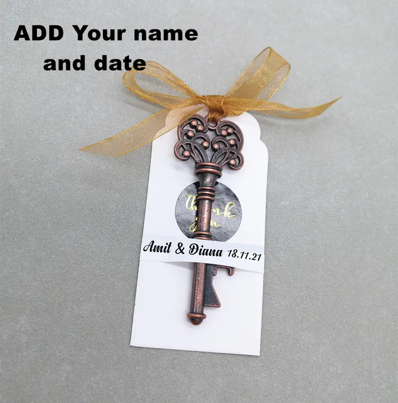 Encanto de llave antigua ornamentada color bronce, elaborado en metal, con una cinta dorada atada en la parte superior, sujeto a una etiqueta blanca, diseñado como recuerdo abridor de botellas para bodas personalizado.