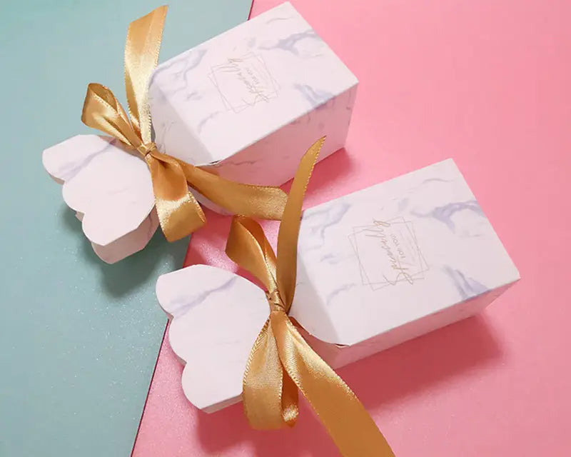 Blumige Hochzeits-Gastgeschenkboxen mit marmoriertem Weiß-Design, gebunden mit goldfarbenen Satinbändern, für eine elegante Geschenkübergabe.