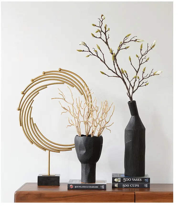 Black textured ceramic vase with vertical ridges and narrow neck holding dried branches with budding flowers as part of gold metal abstract sculpture office desk decor.