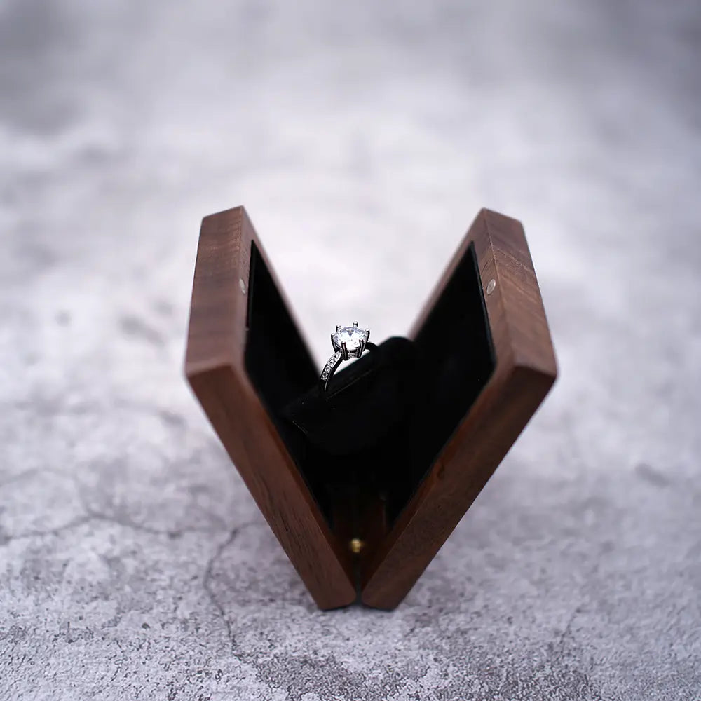 Ein silberner Verlobungsring mit einem zentralen Diamanten und kleineren Diamanten entlang des Bandes, präsentiert in einer geöffneten personalisierten Schmuckring-Schachtel aus Walnussholz zur Hochzeit.