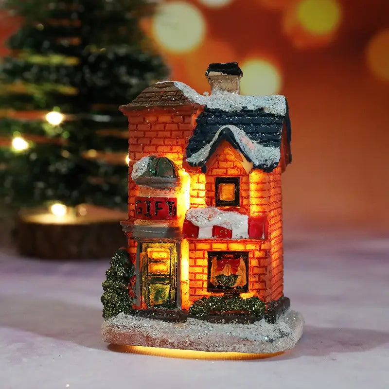 Une petite maison de boutique de cadeaux en brique, miniature, éclairée chaleureusement, avec un toit blanchit par la neige et des fenêtres lumineuses, ornée de buissons verts scintillants et d’un panneau de cadeau, pour un ensemble de maisons de village de Noël, veilleuse à batterie.