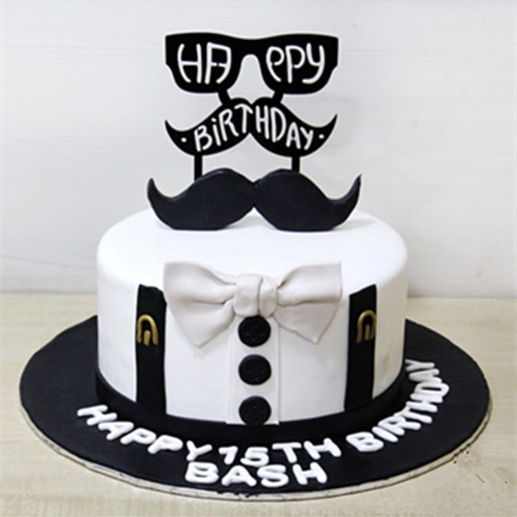 A white frosted birthday cake decorated with a father’s day acrylic cake topper featuring a black mustache and glasses, silver bow tie, and black suspenders with gold accents.