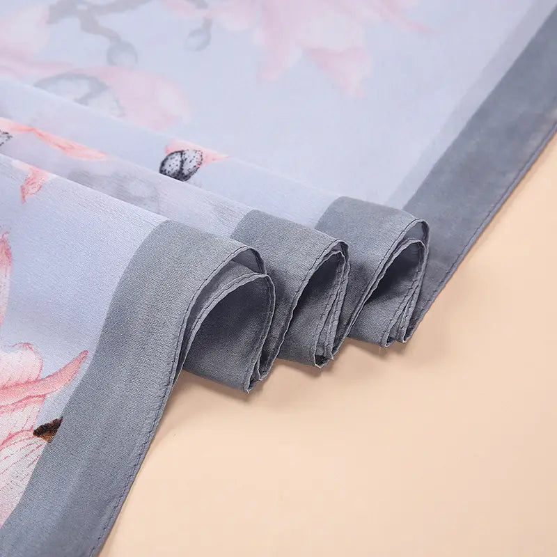 Un châle foulard en soie bleu clair délicat Van Gogh, amandiers en fleurs, avec un motif floral rose pâle et papillons, ainsi qu’une bordure unie grise.