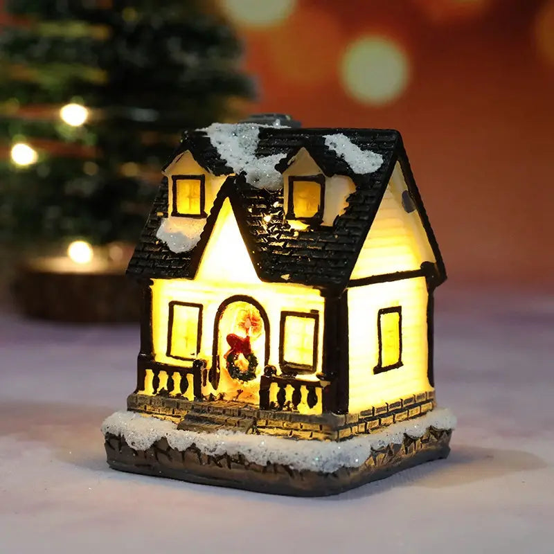 Une figurine miniature de maison du village de Noël à LED, éclairée chaleureusement, avec un toit en ardoises noires et des détails blanchis par la neige, présentant des fenêtres jaunes lumineuses et une couronne festive à la porte d’entrée, veilleuse à batterie.