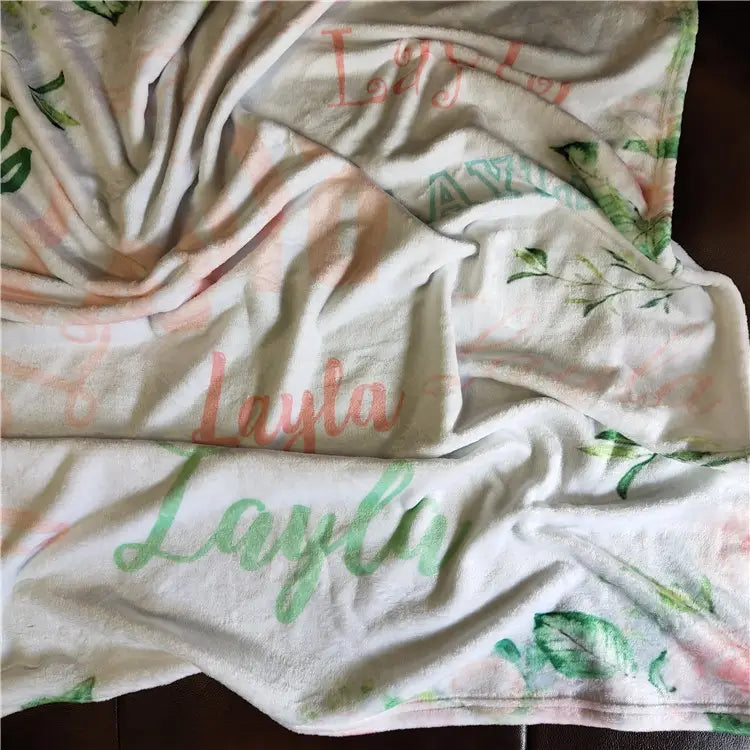 Eine weiche weiße, personalisierte Baby-Wickeldecke aus plüschigem Fleece mit dem Namen Layla, im rosa-grünen Cursive-Stil aufgedruckt, mit zarten Blattmustern.