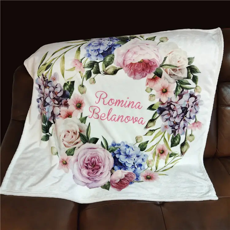 Eine weiße, personalisierte Baby-Wickeldecke aus Fleece mit einer plüschigen Textur und einem detaillierten Design eines floralen Kranzes aus pinken, violetten und blauen Blumen, umrahmt den Namen Romina Belanova in eleganter Schreibschrift.