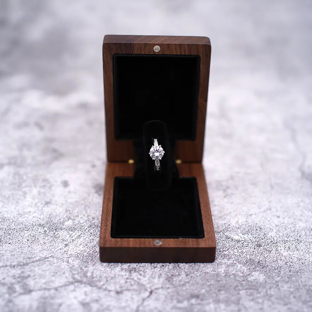 Ein funkelnder Diamant-Verlobungsring mit einem zentralen Stein und kleineren Akzentsteinen, gesetzt in ein schlankes Silberband, präsentiert innerhalb einer geöffneten personalisierten Schmuckring-Schachtel aus Walnussholz zur Hochzeit.