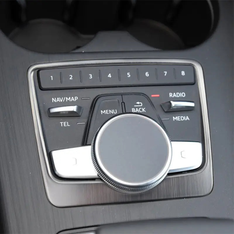 Metallischer Drehregler in Silberoptik mit strukturierter Grifffläche, umgeben von schwarzen Tasten, beschriftet mit „Nav“, „Karten“, „Menü“, „Zurück“, „Radio“ und „Media“ auf der Einfassung des Audi A4 B9 A5 Schalthebel-Wandhalters für den Becherhalter.