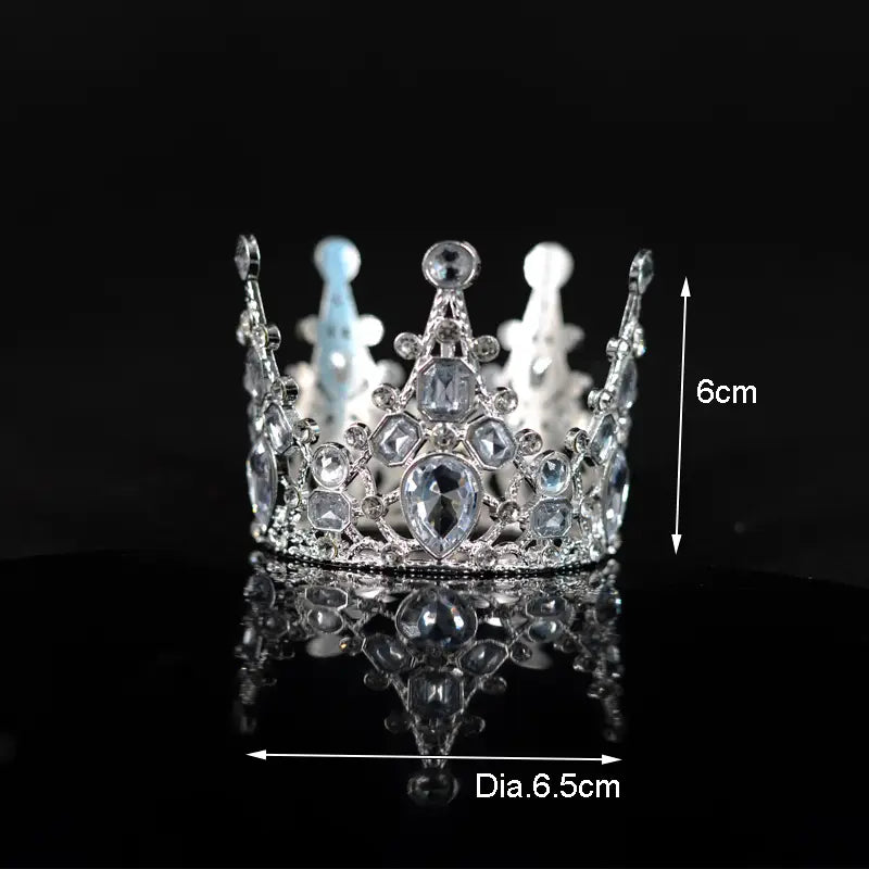Ein funkelnder silberner Mini-Kronen-Tortenaufsatz und eine Haar-Tiara, verziert mit klaren und hellblauen Kristallen, mit einem kunstvollen Design mit einem zentralen, tränenförmigen Stein.