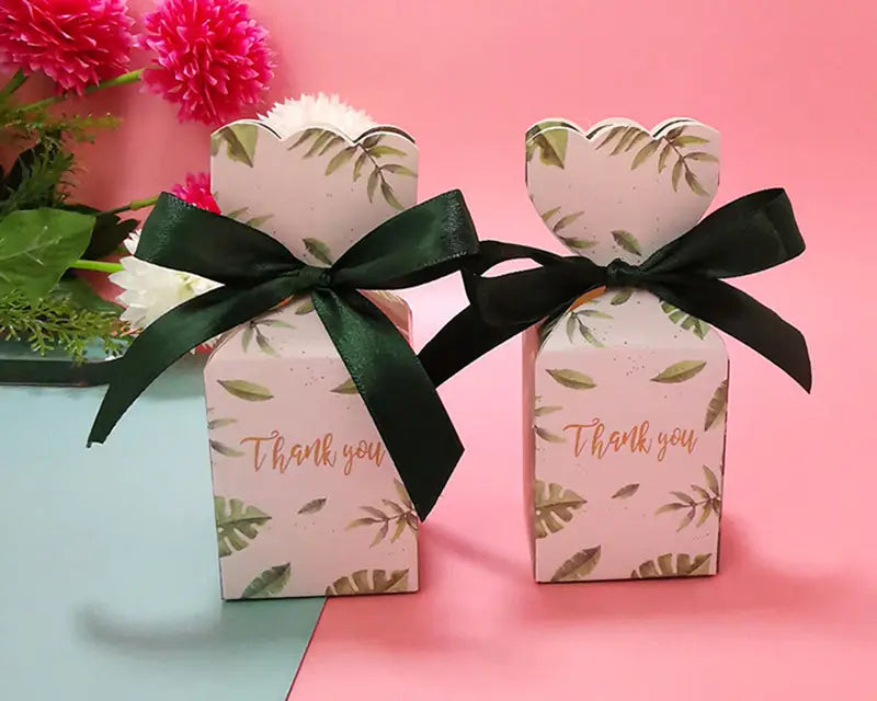 Rosa Papier-Gastgeschenkboxen mit blumigen Mustern, grünen Blattmustern und goldfarbenem „Danke“-Text, gebunden mit schwarzen Satinbändern.