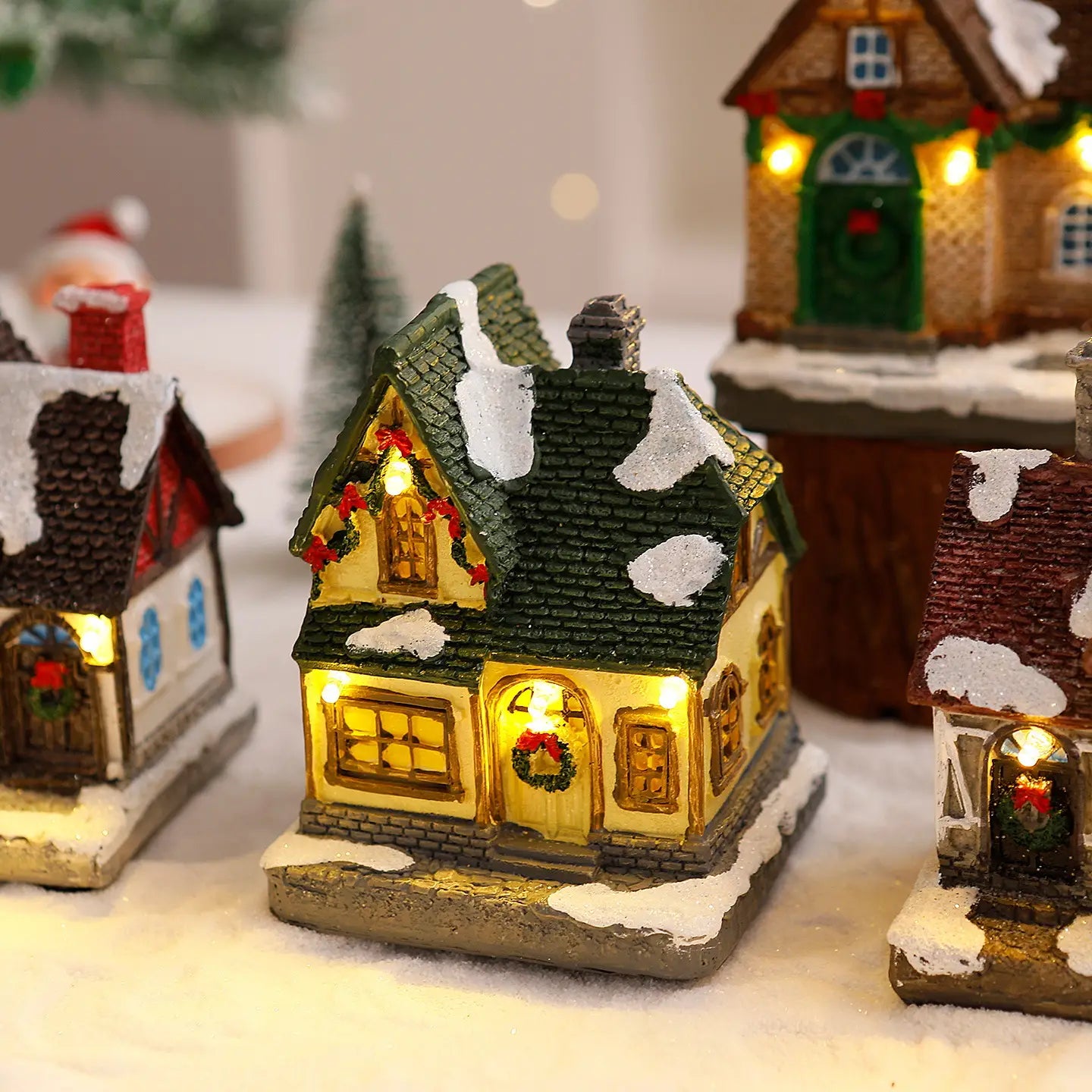Ornement de maison de village de Noël éclairée, allumé chaleureusement, avec un toit vert, des détails comme enneigés et des couronnes festives sur les fenêtres.