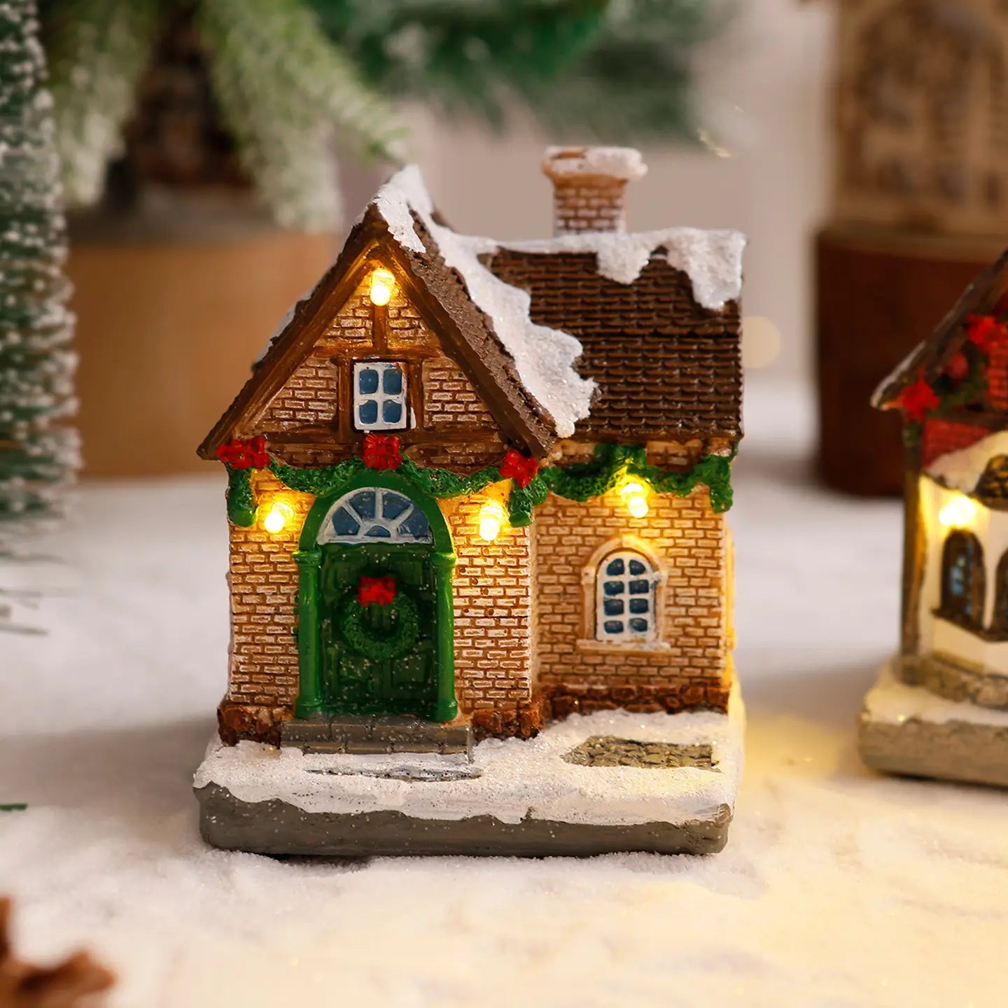 Ornement de maison de village de Noël éclairée présentant une façade miniature en briques brunes, une porte verte cintrée décorée d’une couronne rouge et un toit couvert de neige illuminé par des lumières intérieures chaudes.