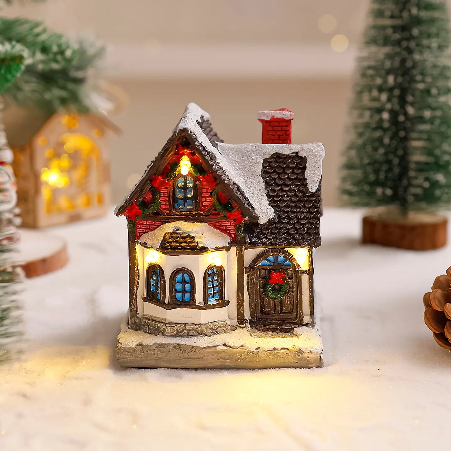 Ornement figurine miniature de maison de village de Noël éclairée, avec un toit couvert de neige, des murs en briques rouges et des fenêtres lumineuses.