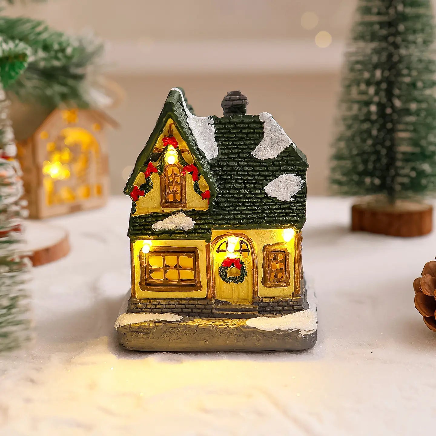 Ornement de maison de village de Noël éclairée présentant une maison miniature illuminée avec un toit vert, des murs jaunes, et des couronnes rouges festives sur les fenêtres et la porte.