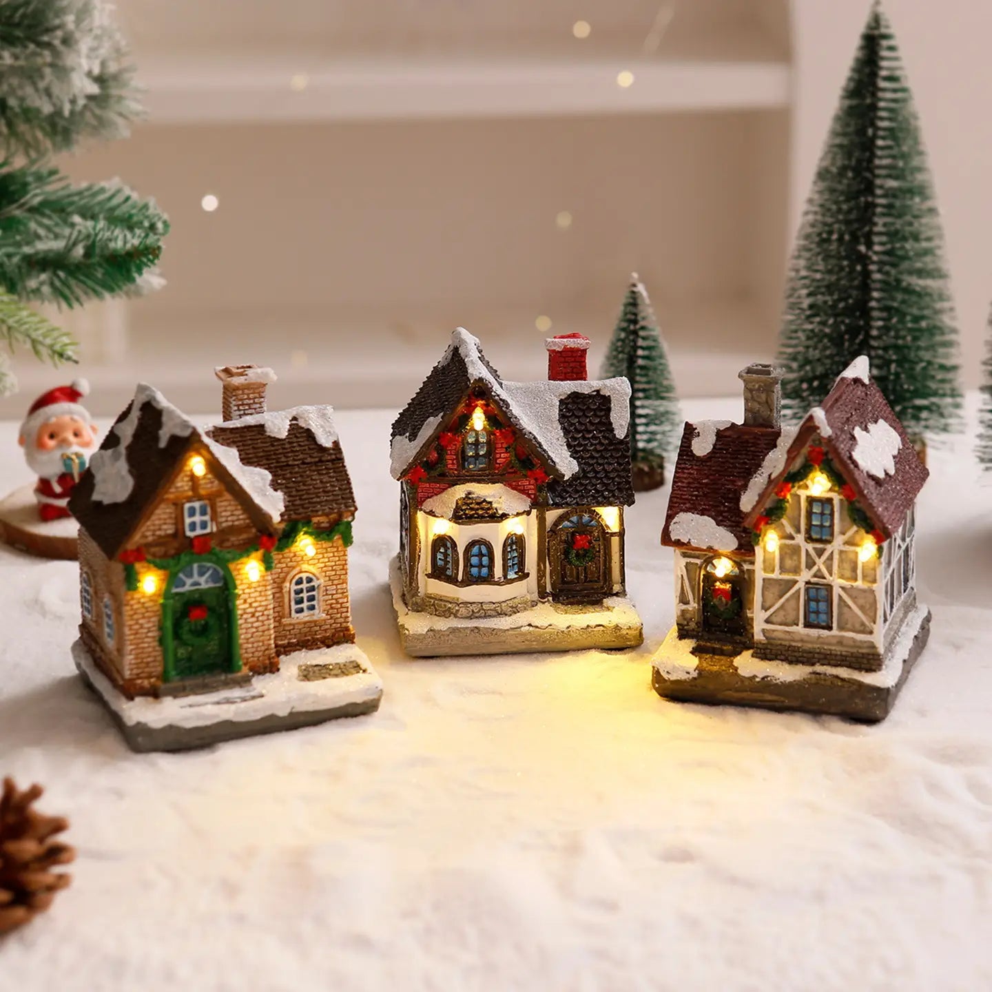 Trois ornements de maison de village de Noël miniature illuminés et éclairés, avec des toits couverts de neige, des lumières festives brillantes et une construction en résine détaillée évoquant l’architecture européenne traditionnelle