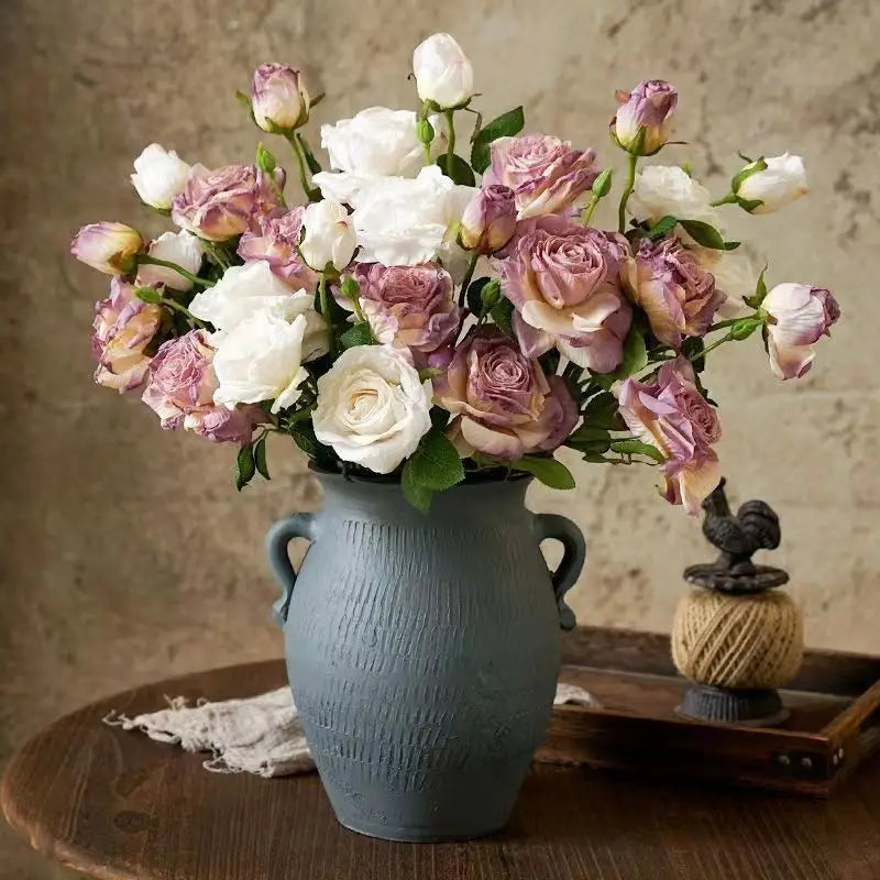 Eine matte blaue Keramikvase mit doppelten Henkeln und einer strukturierten Oberfläche hält einen verwelkten Stielrand-Rosenstrauß mit weißen und lavendelfarbenen Blumen in frontaler Ansicht.