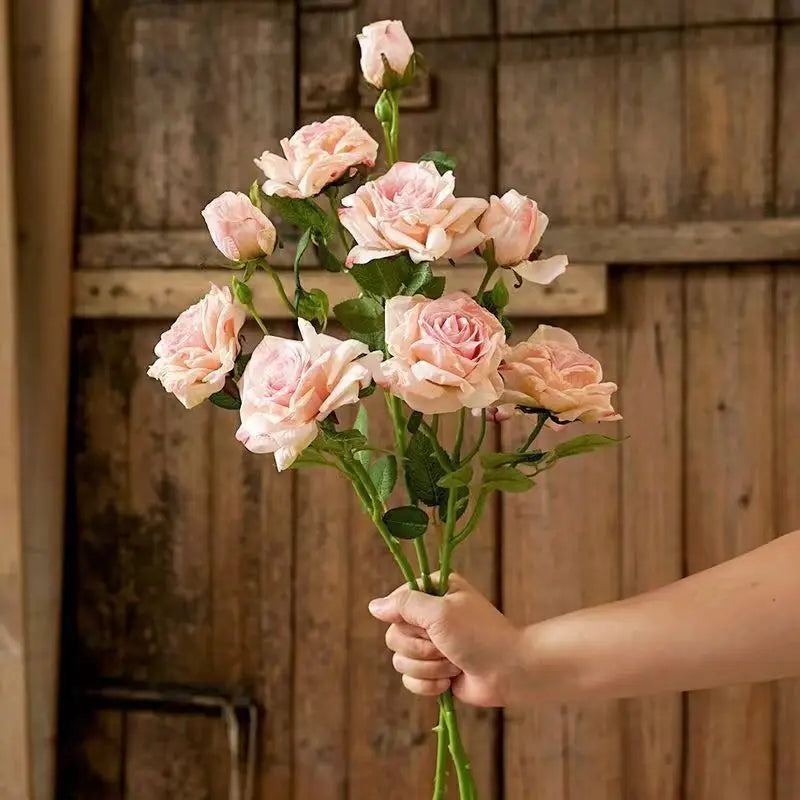 Ein Strauß aus zarten rosa verwelkten Stielrand-Rosen mit grünen Stielen und Blättern, in einer Hand gehalten vor einem rustikalen Holz-Hintergrund.