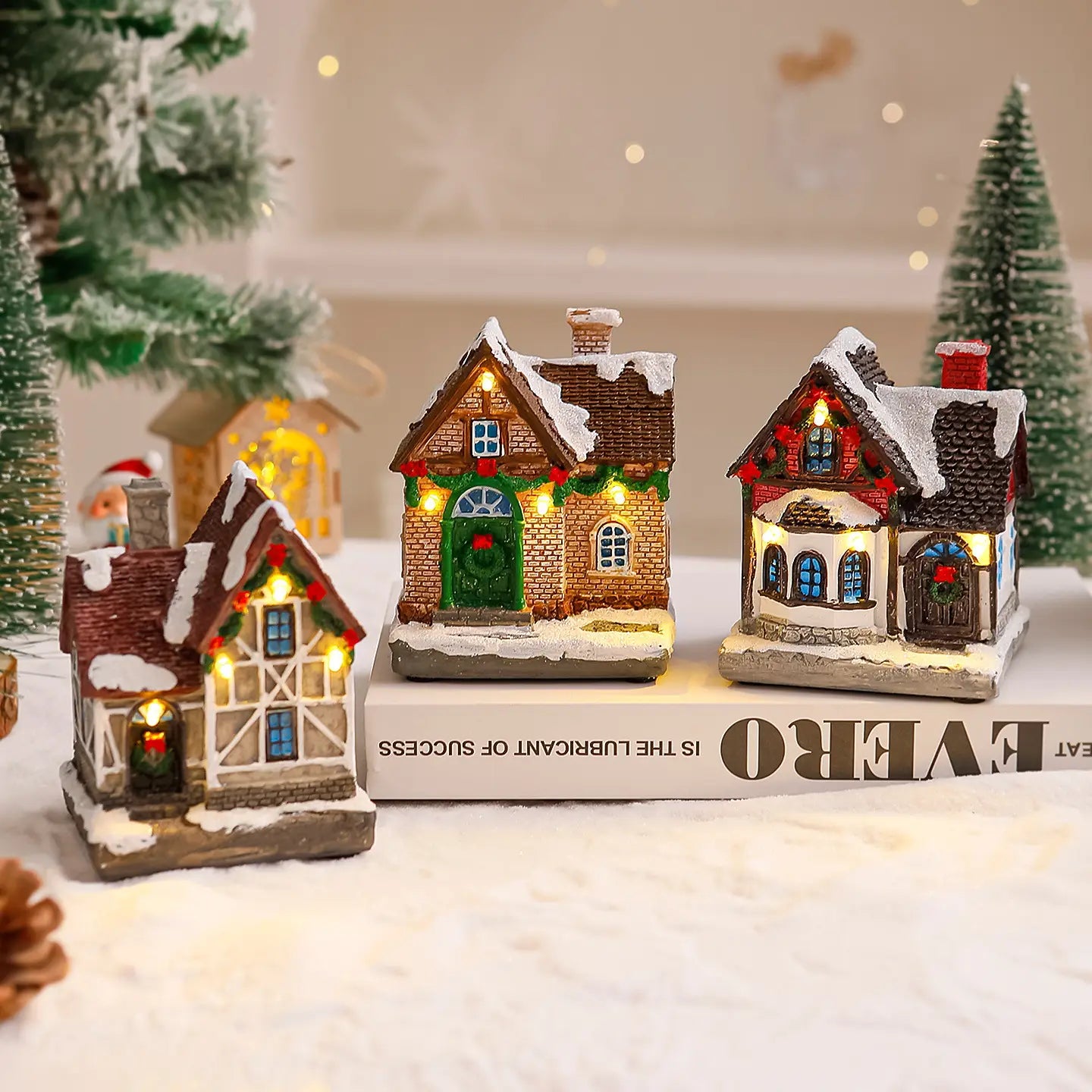 Trois ornements de maison de village de Noël miniature éclairée avec des fenêtres et des portes illuminées, des toits couverts de neige, une maçonnerie détaillée aux tons terreux, parfaits pour la décoration festive des fêtes.